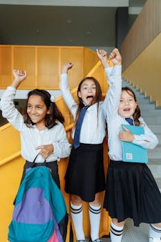 Three cheerful schoolgirls in uniforms celebrating on a staircase indoors.